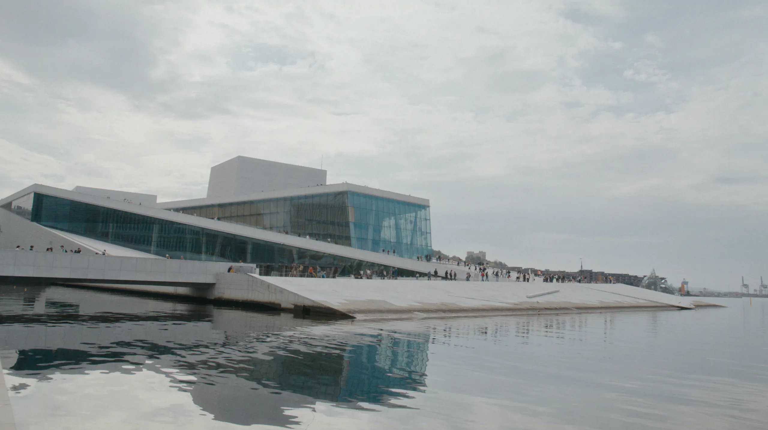 Modern waterfront building with angular white terraces and glass facade beside calm water, with reflections and a cloudy sky
