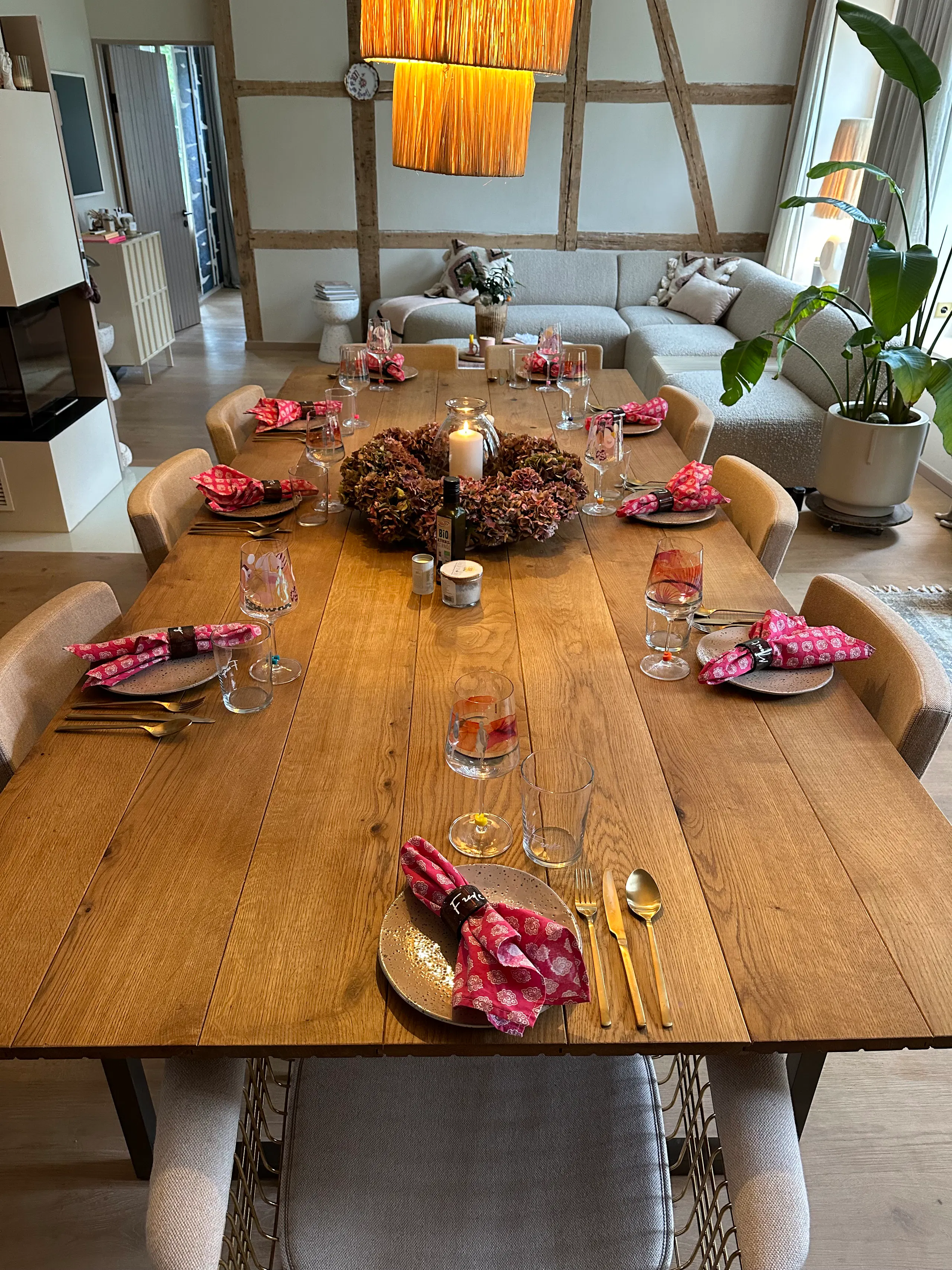 Wooden dining table set for six with plates, glasses, gold cutlery, and pink patterned napkins, centerpiece with candles and dried flowers, modern living room with beige sectional sofa and large potted plant in the background